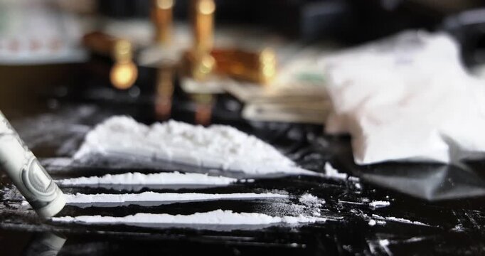 Snorting a line of cocaine with a rolled dollar bill on black background, slow motion. Illegal drugs and cash with gun and bullets on table - crime scene. Criminal problems. Illegal selling