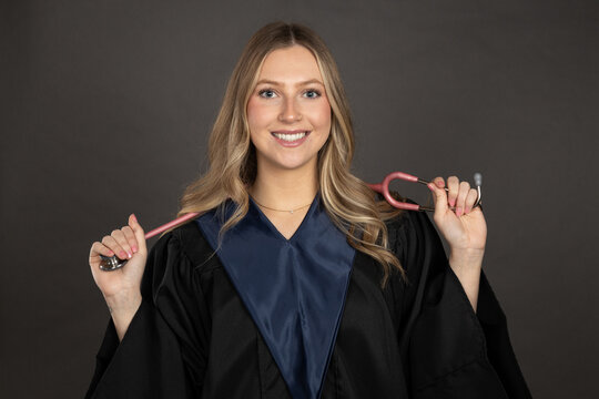 portrait of a young woman in graduation gown holding pink stethoscope, isolated on grey background