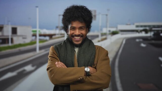 Smiling man with curly hair and arms crossed stands confidently on a quiet highway wearing a warm scarf and brown coat.