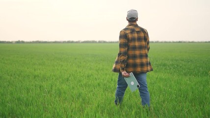 farmer with tablet in the field. business lifestyle finance nature and people concept. a farmer in a hat and plaid shirt walks through a green field with a tablet in his hands - Powered by Adobe