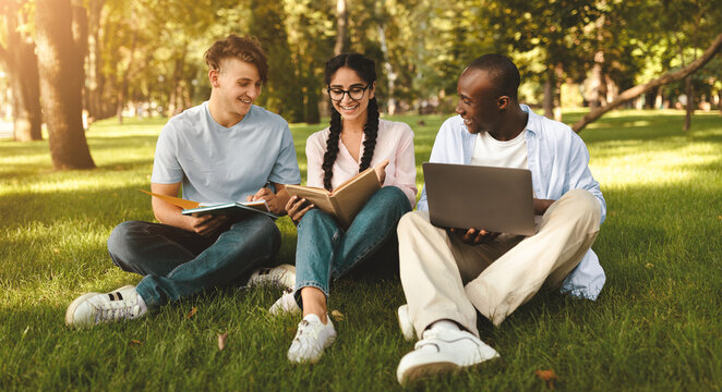 Group of diverse students friends learning, reading books, using laptop and discussing something in university campus park, sitting on grass outside