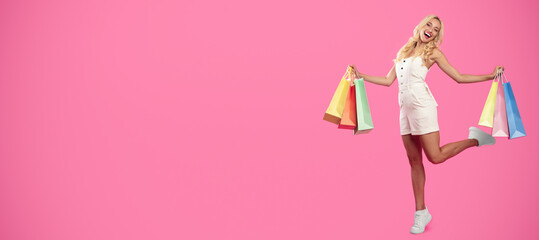 Female Shopping Concept. Full body length shot of excited stylish blonde lady walking and posing with raised leg holding shopper bags from mall, happy about purchase or gifts, pink studio background