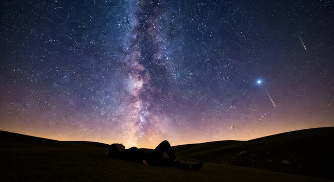 Woman lying down on a hill looking up at the milky way and shooting stars, concept of dreaming and stargazing.