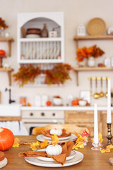Dinning table is set for dinner with white and orange pumpkins, fall leaves, and candlestick on kitchen background. Home decor for Halloween, Thanksgiving day and fall holidays.