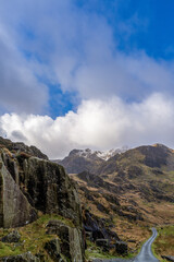 Dramatic mountain landscape in Wales with rocky cliffs, winding trail, and snow-dusted peaks beneath a bright cloudy blue sky