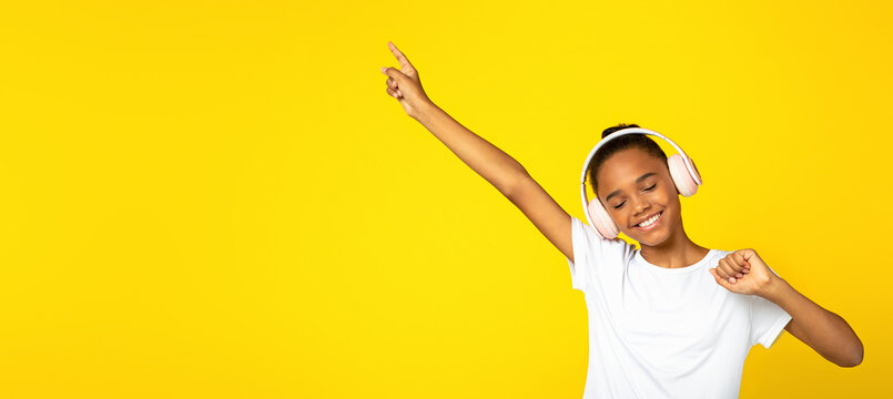 Happy teenager african american girl pupil in white t-shirt and wireless headphones listens music and dances, isolated on yellow background, copy space. Rest, relax in free time, entertainment alone