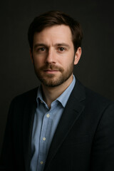 Professional studio portrait of a confident young businessman with a beard, wearing a suit and light blue shirt, captured in soft, natural lighting against a dark background.