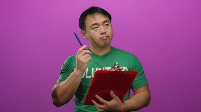 Young man standing with clipboard wearing green shirt against pink wall isolated background thinking thoughtful.