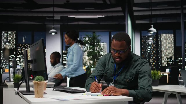African american advisor extracting data by hand on a notebook late at night, drafting highlights from finance reports for the business administration. Notetaking supports goal setting. Camera A.