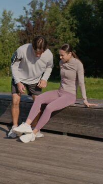 Vertical shot of active couple jogging along wooden deck in park while female athlete sprained her ankle and sitting down on bench, guy putting her leg on his knees and trying to help
