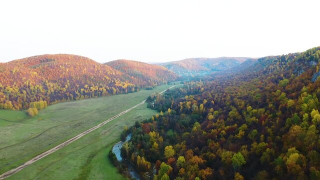 aerial perspective captures the vibrant autumn colors of the Kyzyltash Mountain Range in the South Ural region of Russia. The footage showcases a winding river flowing through a lush green valley, fla