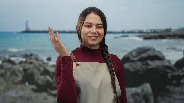 Woman in apron gesturing near seaside with rocks and ocean in background, embracing nature while smiling in outdoor setting on a sunny day at the beach.