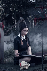 A young woman dressed in black, holding a grim reaper's scythe and a black magic wand, stands in the middle of a graveyard surrounded by graves, smoke, and human skulls. Halloween costume idea.