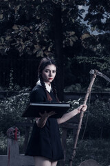 A young woman dressed in black, holding a grim reaper's scythe and a black magic wand, stands in the middle of a graveyard surrounded by graves, smoke, and human skulls. Halloween costume idea.
