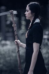 A young woman dressed in black, holding a grim reaper's scythe and a black magic wand, stands in the middle of a graveyard surrounded by graves, smoke, and human skulls. Halloween costume idea.