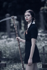 A young woman dressed in black, holding a grim reaper's scythe and a black magic wand, stands in the middle of a graveyard surrounded by graves, smoke, and human skulls. Halloween costume idea.