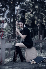 A young woman dressed in black, holding a grim reaper's scythe and a black magic wand, stands in the middle of a graveyard surrounded by graves, smoke, and human skulls. Halloween costume idea.