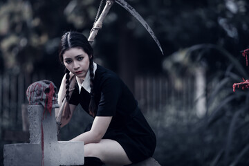 A young woman dressed in black, holding a grim reaper's scythe and a black magic wand, stands in the middle of a graveyard surrounded by graves, smoke, and human skulls. Halloween costume idea.