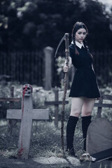 A young woman dressed in black, holding a grim reaper's scythe and a black magic wand, stands in the middle of a graveyard surrounded by graves, smoke, and human skulls. Halloween costume idea.
