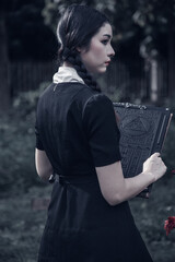 A young woman dressed in black, holding a grim reaper's scythe and a black magic wand, stands in the middle of a graveyard surrounded by graves, smoke, and human skulls. Halloween costume idea.