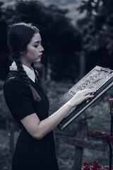 A young woman dressed in black, holding a grim reaper's scythe and a black magic wand, stands in the middle of a graveyard surrounded by graves, smoke, and human skulls. Halloween costume idea.