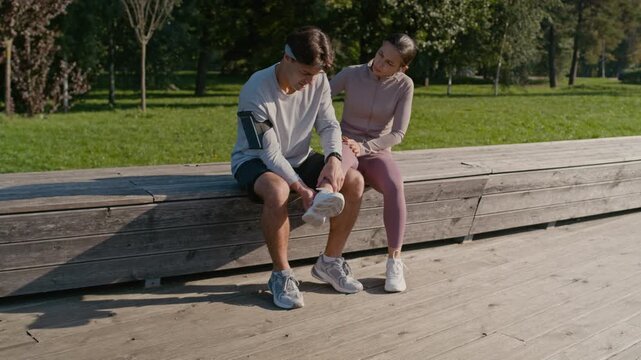 Young sportsman sitting on wooden bench in park next to his girlfriend with sprained ankle and trying to help her while examining leg of female athlete