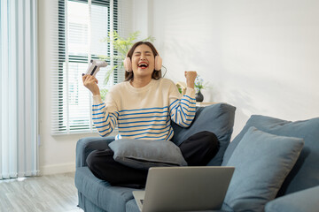 A young woman wearing headphones enjoys playing a video game on her laptop while sitting on a sofa at home, expressing excitement and joy during the gameplay.