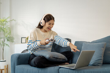 A young woman wearing headphones enjoys playing a video game on her laptop while sitting on a sofa at home, expressing excitement and joy during the gameplay.