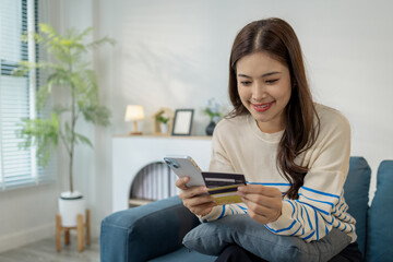 A young woman is sitting on a sofa holding a smartphone and credit card, smiling as she makes an online transaction. She looks relaxed and confident while shopping at home.