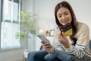 A young woman is sitting on a sofa holding a smartphone and credit card, smiling as she makes an online transaction. She looks relaxed and confident while shopping at home.