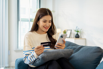 A young woman is sitting on a sofa holding a smartphone and credit card, smiling as she makes an online transaction. She looks relaxed and confident while shopping at home.