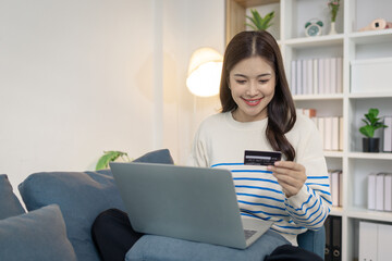 A young woman sits comfortably on a sofa using a laptop while holding a credit card, appearing to make an online payment. She smiles with satisfaction, reflecting ease and convenience.