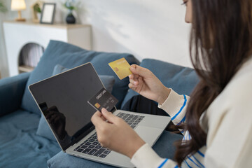A young woman sits comfortably on a sofa using a laptop while holding a credit card, appearing to make an online payment. She smiles with satisfaction, reflecting ease and convenience.