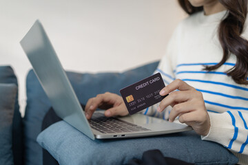 A young woman sits comfortably on a sofa using a laptop while holding a credit card, appearing to make an online payment. She smiles with satisfaction, reflecting ease and convenience.