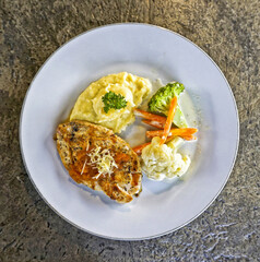 Top view of grilled chicken steak served with mashed potatoes, broccoli, carrot, and cauliflower on white plate. Healthy and delicious western meal concept for restaurant, catering, or diet food promo