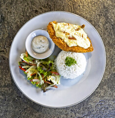 Top view of crispy chicken cordon bleu served with white rice, fresh salad, and creamy sauce on a white plate. Perfect for restaurant menu, catering, or Asian fusion food promotion