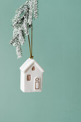 Minimal winter white ceramic house ornament hanging on snow-covered fir branch on green colored background. Christmas decoration symbolizing home, warmth and holiday coziness. New Year Christmas decor