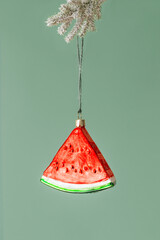 Minimal creative Christmas holidays concept. Red green slice of watermelon hanging on xmas snowy fir tree branch on green. Winter seasonal still life with Christmas decoration, New Year modern design