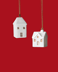 Minimal winter composition with two white ceramic house ornament hanging on red background. Simple Christmas decoration symbolizing home, warmth and holiday coziness. New Year colored Christmas decor.