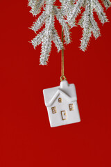 White small house ornament hanging on snowy fir branch on bright red background. Side view New Year cute toy. Concept of real estate, new home, family comfort and holiday mood, winter seasonal design.