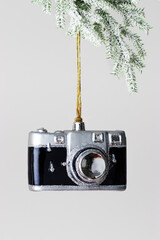 Minimal winter composition with toy camera Christmas ornament hanging from snowy fir branch on light gray background, creative holiday concept connecting photography, travel memory and New Year.