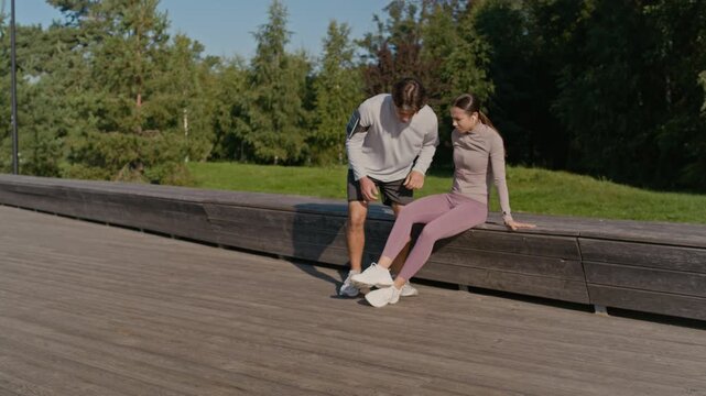 Young active couple jogging along wooden deck in park on sunny day while female athlete sprained her ankle and sitting down on bench, guy putting her leg on his knees and trying to help