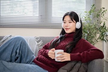 Asian woman wearing headphones holding coffee mug cup while sitting leaning on sofa in a living room