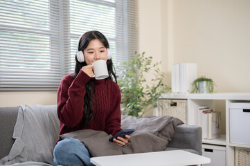 Asian woman wearings headphones holding phone drinking coffee while sitting on sofa in a living room