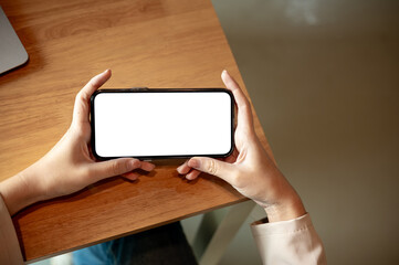 Woman hands holding playing blank screen smartphone horizontally on wooden table in cafe or library.