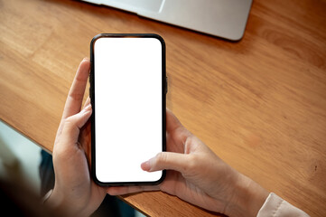 Close up of woman hand holding typing on blank screen smartphone on wooden table in cafe or library.