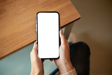 Close up of woman holding blank screen smartphone with both hands on wooden table in cafe or library