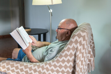 An elderly man enjoys a quiet moment reading a book while sitting in a comfortable chair. Soft light from a nearby lamp adds warmth to the relaxing atmosphere.