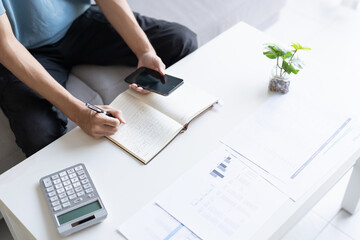 Asian Man Taking Notes on Finances, Budget, Household Expenses, and Tax Calculations in Notebook at...