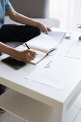 Asian Man Taking Notes on Finances, Budget, Household Expenses, and Tax Calculations in Notebook at Home.
